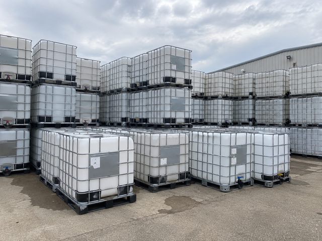 Rows of used caged IBC totes stacked two high on a concrete pad under an overcast sky