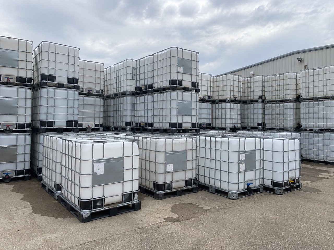 Rows of used caged IBC totes stacked two high on a concrete pad under an overcast sky