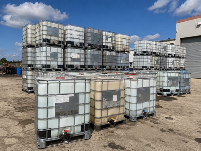 Mixed-condition IBC totes in an outdoor yard under a blue sky, including one heavily stained amber unit