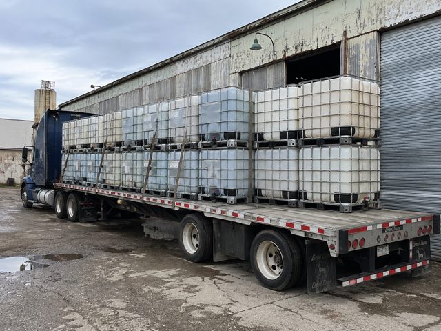 A blue tractor unit with a flatbed trailer loaded with strapped IBC totes beside a warehouse roller door
