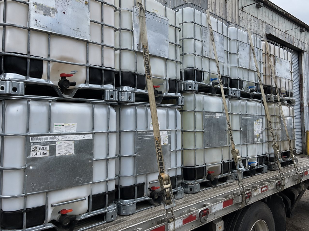 Close view of IBC totes strapped in two tiers on a flatbed trailer, showing 275 gallon labels and ratchet straps